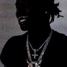 The album cover for A Great Chaos, revealed on October 6th, 2023. It features a grainy, poorly lit photo of Ken Carson wearing a black shirt, multiple diamond chains, and a single earring as he smiles in front of a gray backdrop.