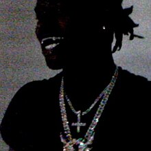 The album cover for 'A Great Chaos', revealed on October 6th, 2023. It features a grainy, poorly lit photo of Ken Carson wearing a black shirt, multiple diamond chains, and a single earring as he smiles in front of a gray backdrop.