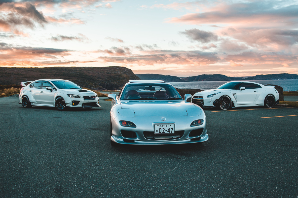 Three Beautiful Japanese vehicles starting with the Subaru WRX Sti on the left a newer Nissan GT-R on the right and in the center a Mazda RX-7 known for its popup headlights, aggressive frontend, and unique rotary engine rarely if not ever found in modern vehicles 