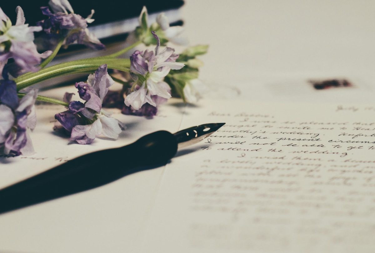 Purple flowers on paper, with cursive writing in ink. A fountain pen lies on top of the paper, alongside a black-covered book in the background.