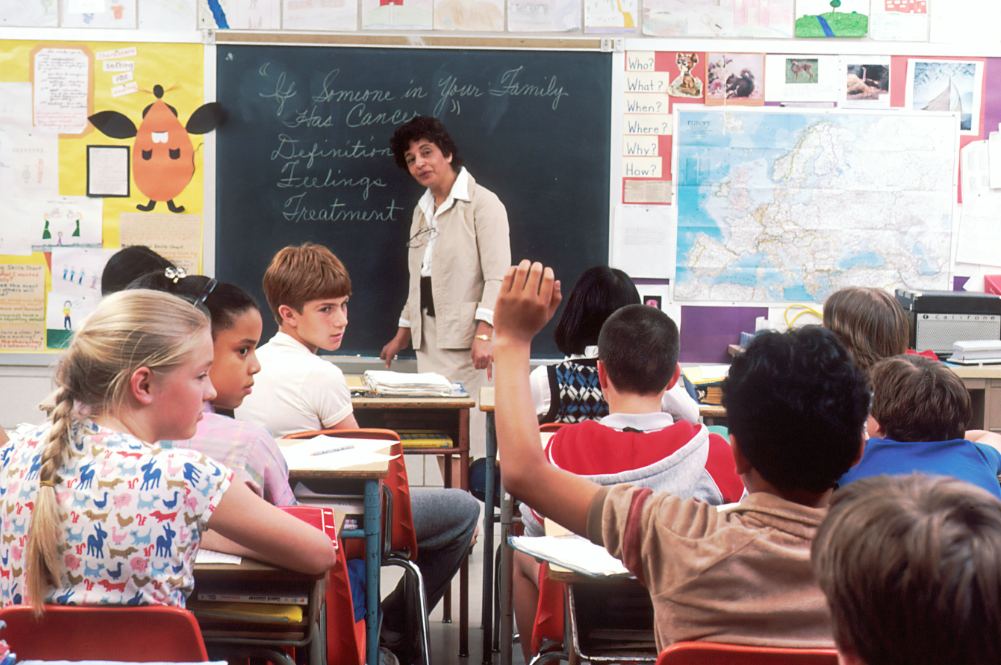 A female teacher standing in front of children in a classroom. In the back of a classroom, are children about 11 years old with a female teacher talking about the subject.